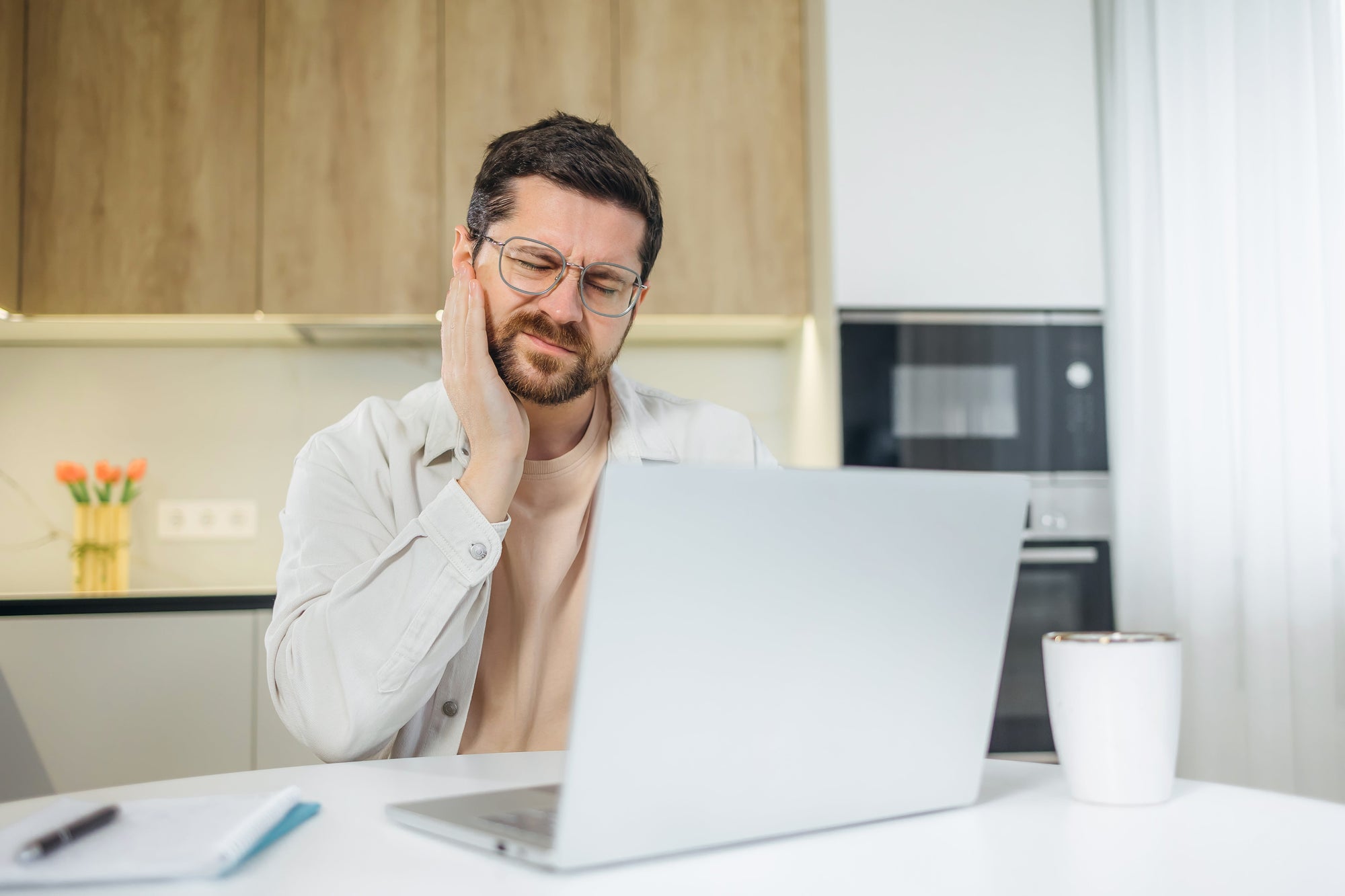 Foto eines Mannes der am Schreibtisch vor seinem Laptop sitzt. Er fasst sich mit der Hand an den Kiefer und hat einen schmerzverzehrten Gesichtausdruck. 